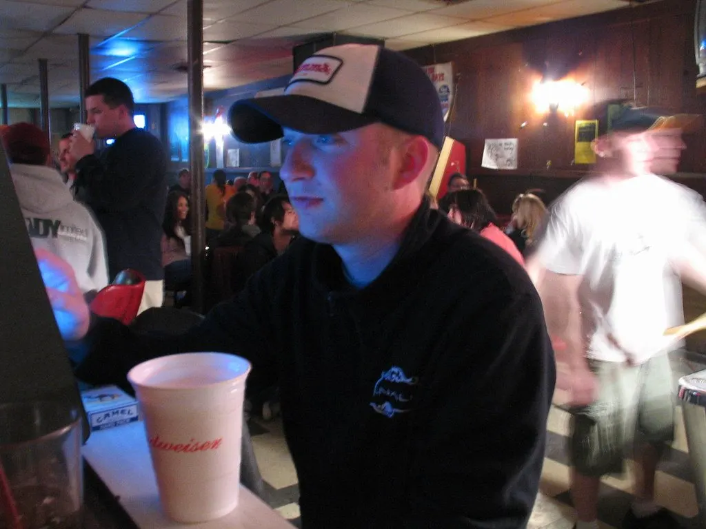 A man in a cap playing a countertop touchscreen game at a crowded bar in the 2000s