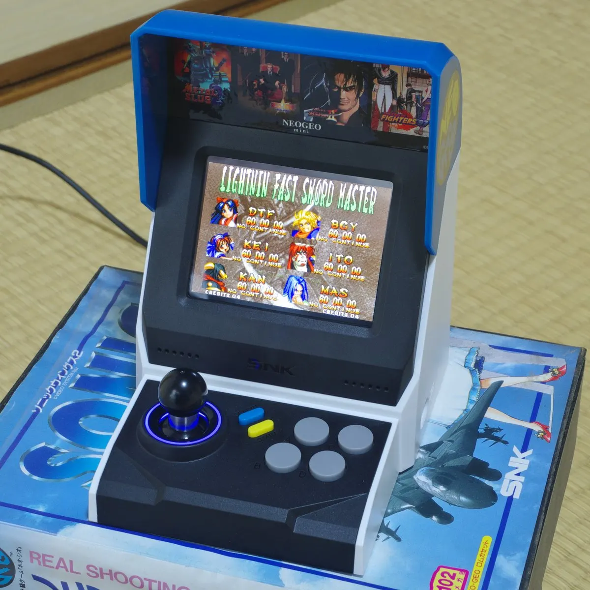 A palm-sized Neo Geo Mini desktop arcade cabinet with a small screen, joystick and four buttons
