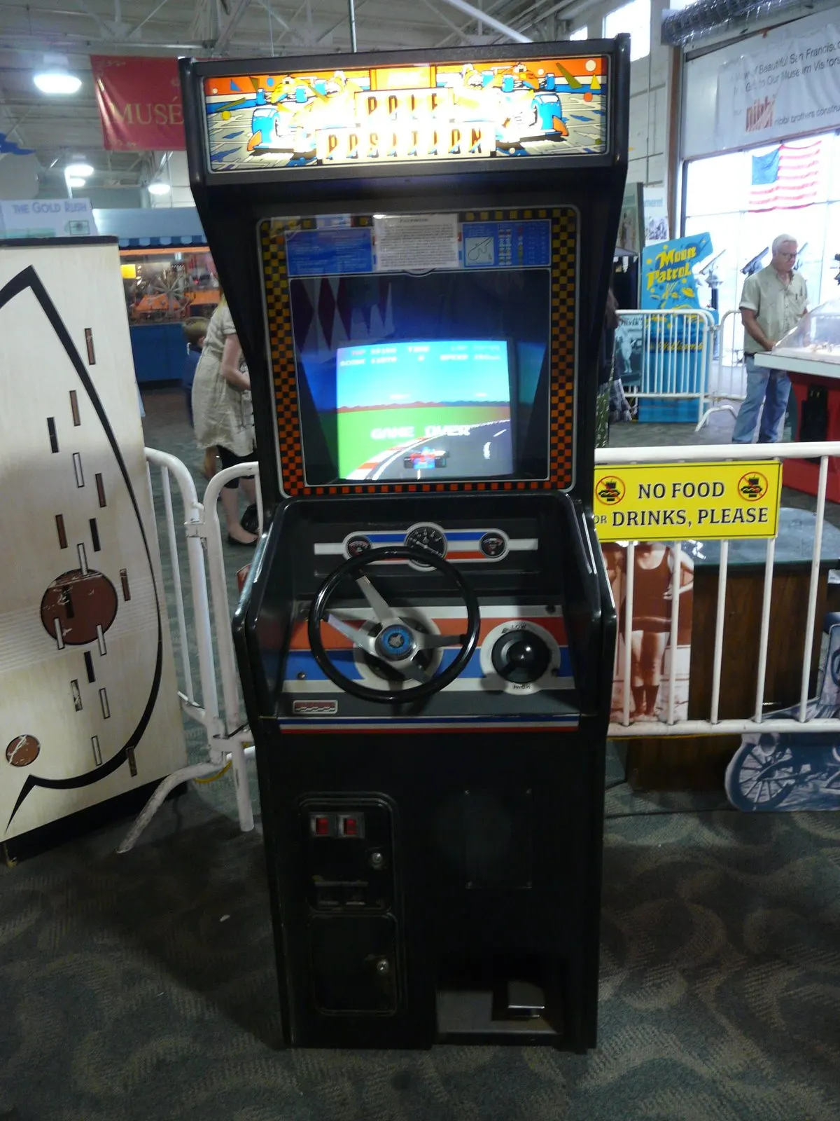 A full-size 1980s upright arcade cabinet with a steering wheel on the control panel, standing in a museum hall