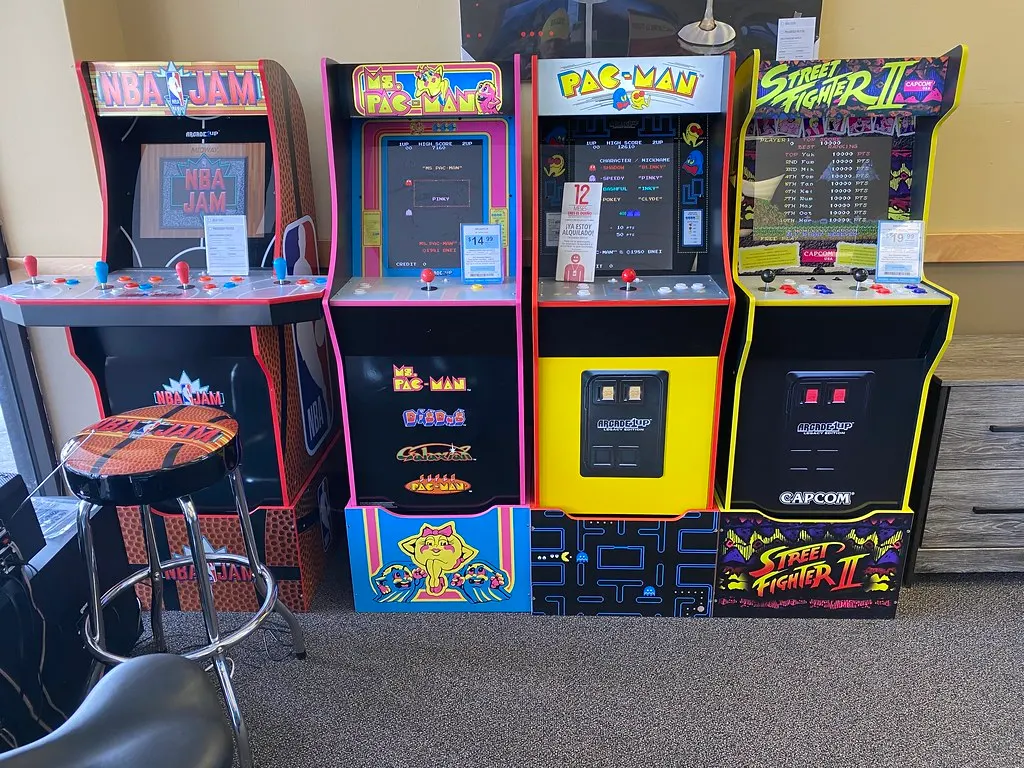 Four home arcade cabinets standing in a row along the wall of a carpeted room, with a bar stool in front